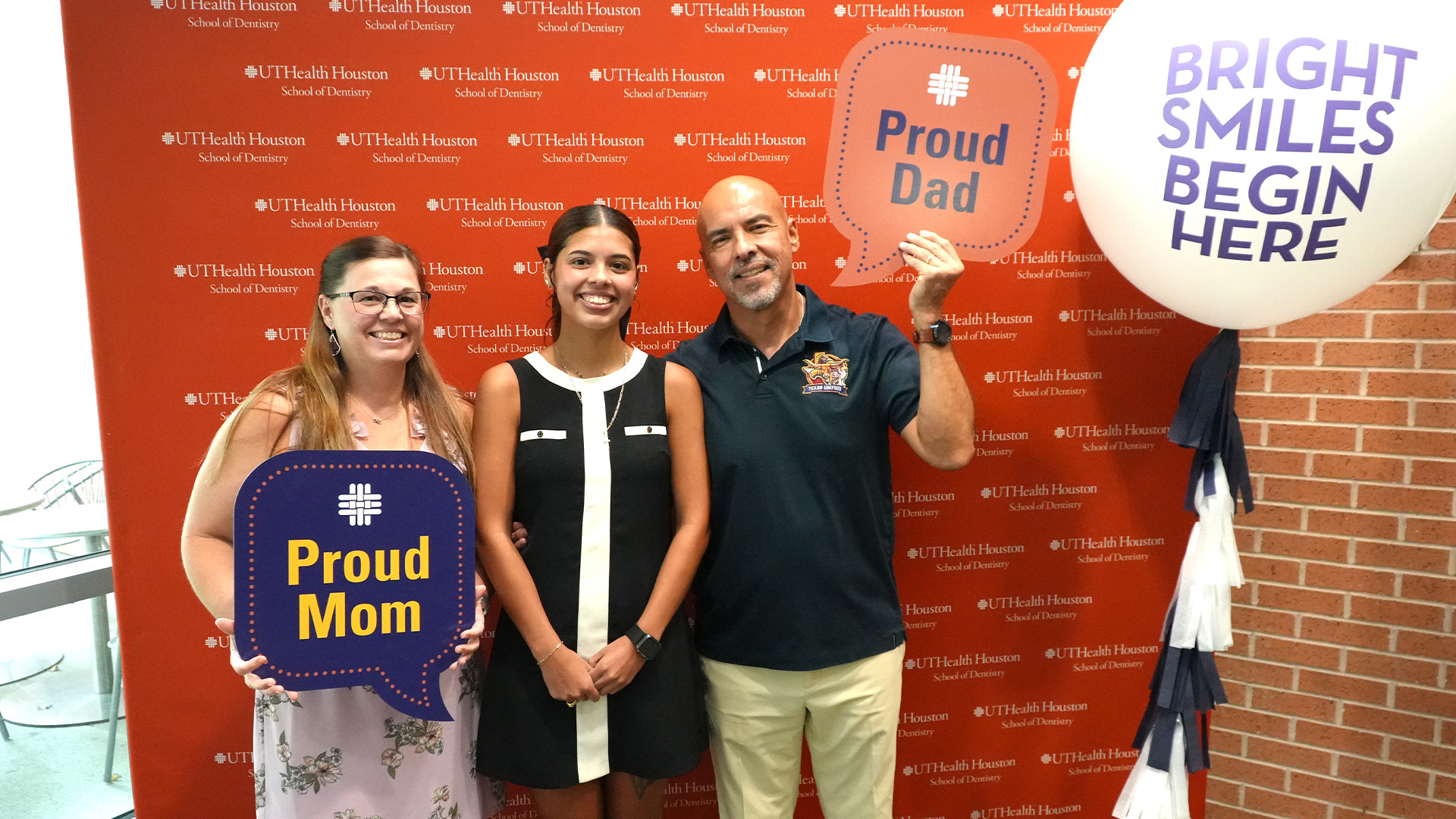 Three people stand in front of a UTHealth Houston School of Dentistry backdrop, with two holding signs that read Proud Mom and Proud Dad beside a large balloon that says Bright Smiles Begin Here.