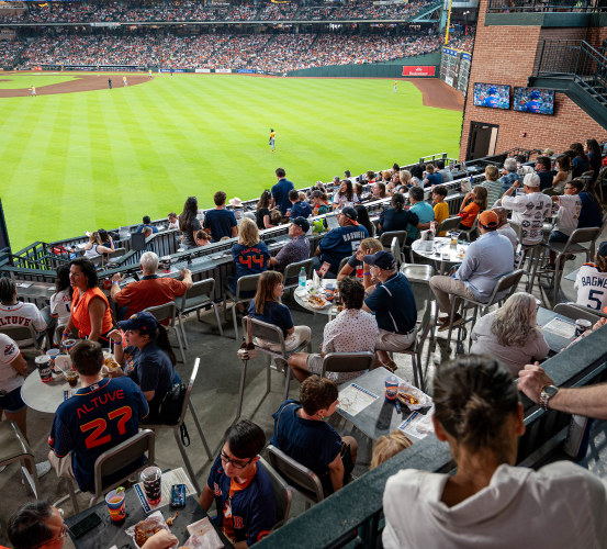 Fans wearing Houston Astros team apparel seated in the center field deck enjoy food and drinks while watching a baseball game at Daikin Park.