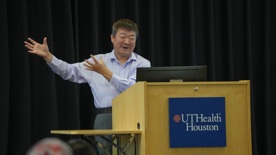 Person giving a presentation at a podium with a UTHealth Houston sign, gesturing with both hands.