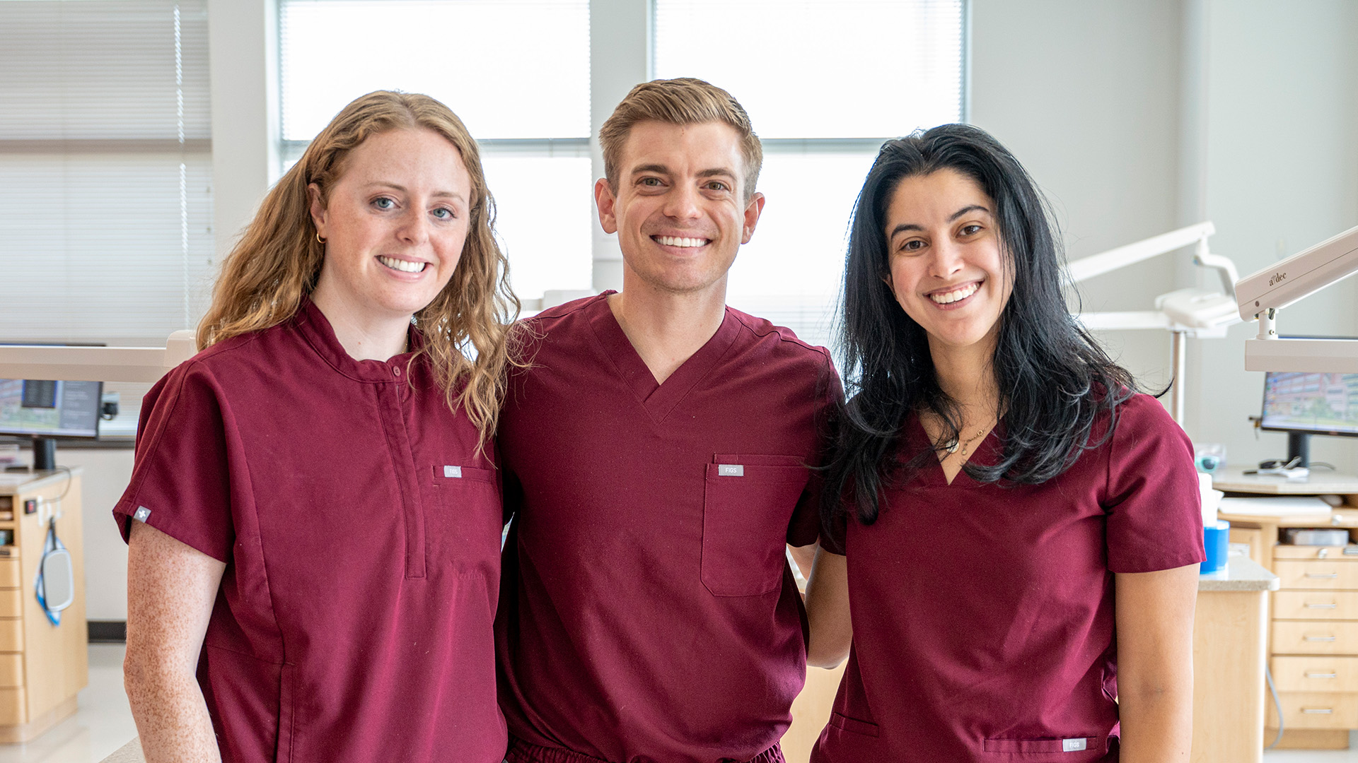 Three individuals in matching maroon scrubs stand together in a dental operatory with clinical equipment and cabinetry behind them.