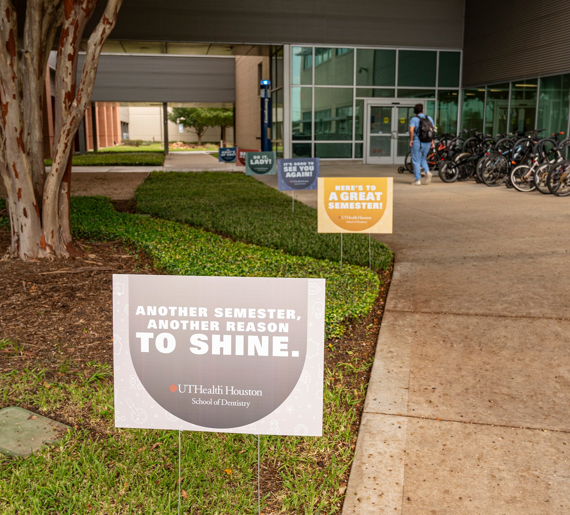 Yard signs with motivational messages line the sidewalk outside UTHealth Houston School of Dentistry. A person in blue scrubs walks along a bike rack toward the building entrance.