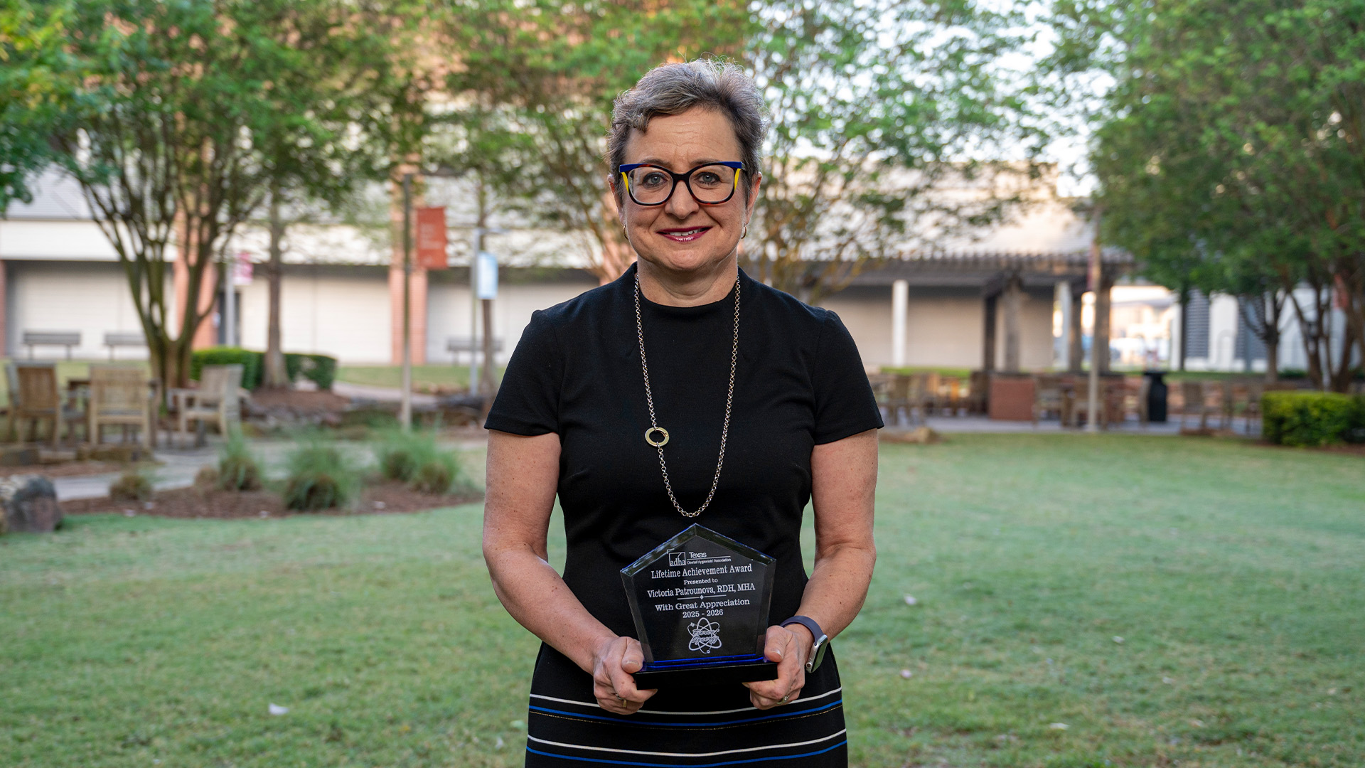 A woman wearing glasses and a black shirt stands outdoors in a landscaped area holding a clear award plaque.