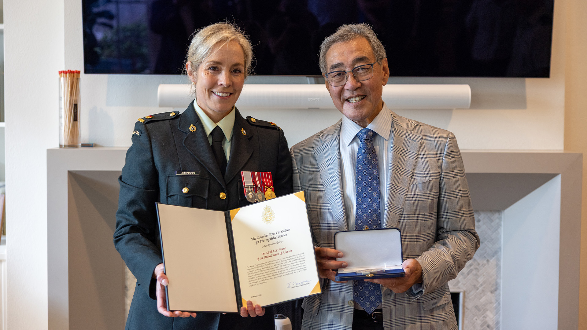 Two individuals pose indoors during a formal ceremony, displaying an open certificate and a plaque.