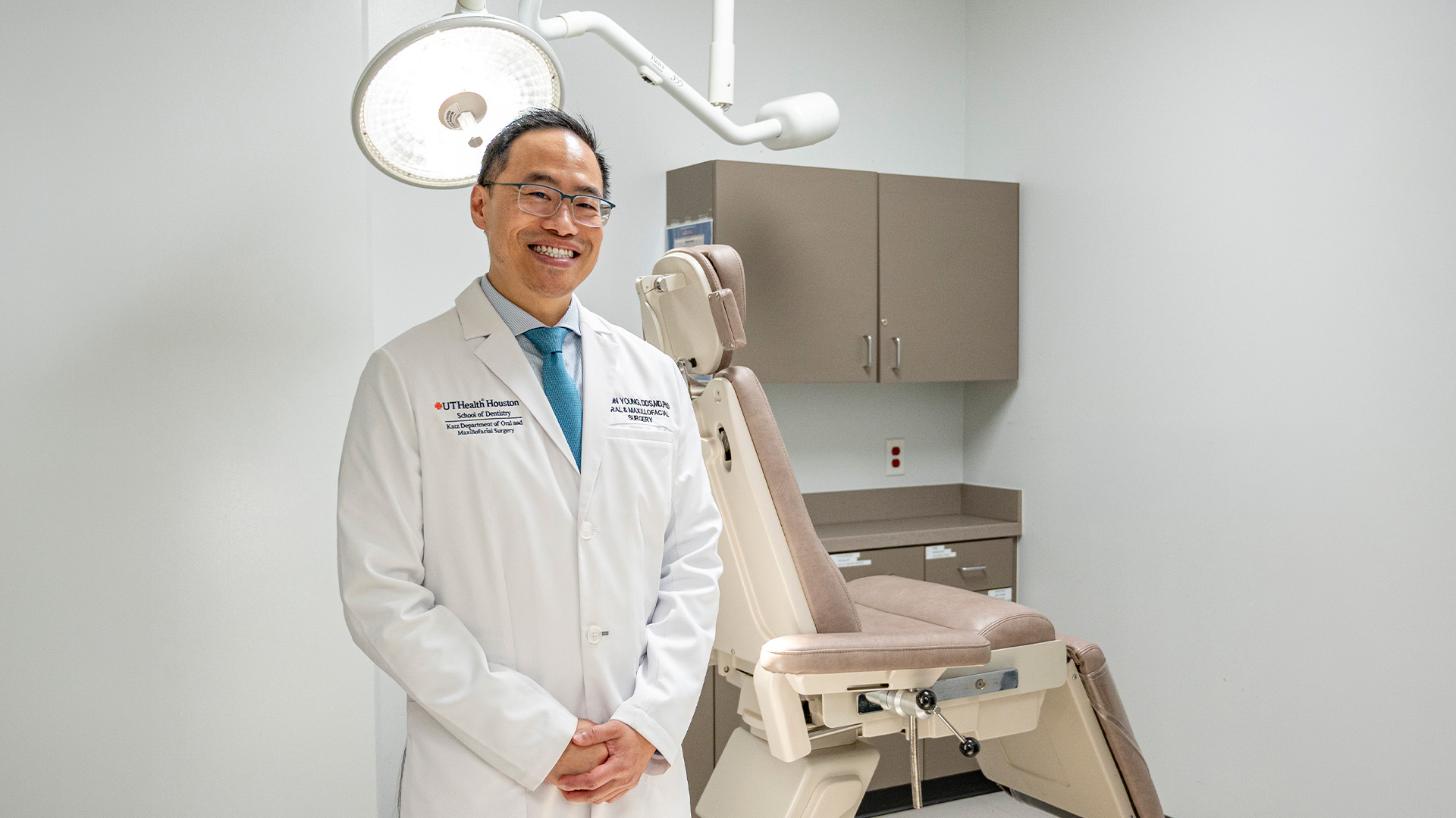 A person in a white UTHealth Houston School of Dentistry coat stands in a clinical exam room beside a beige dental chair, overhead surgical light, and cabinets.