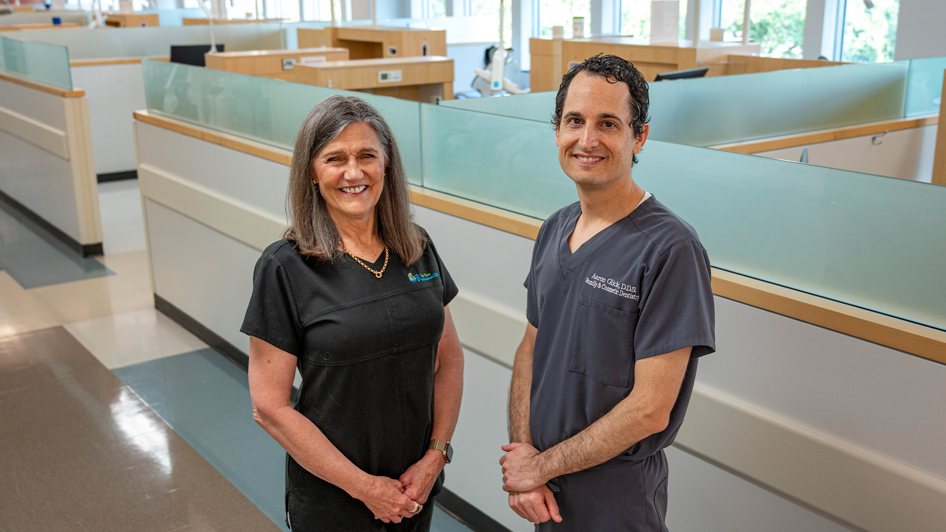 Two dental professionals stand inside a modern clinic with frosted glass partitions, dental workstations, and natural light, wearing black and gray embroidered scrubs.