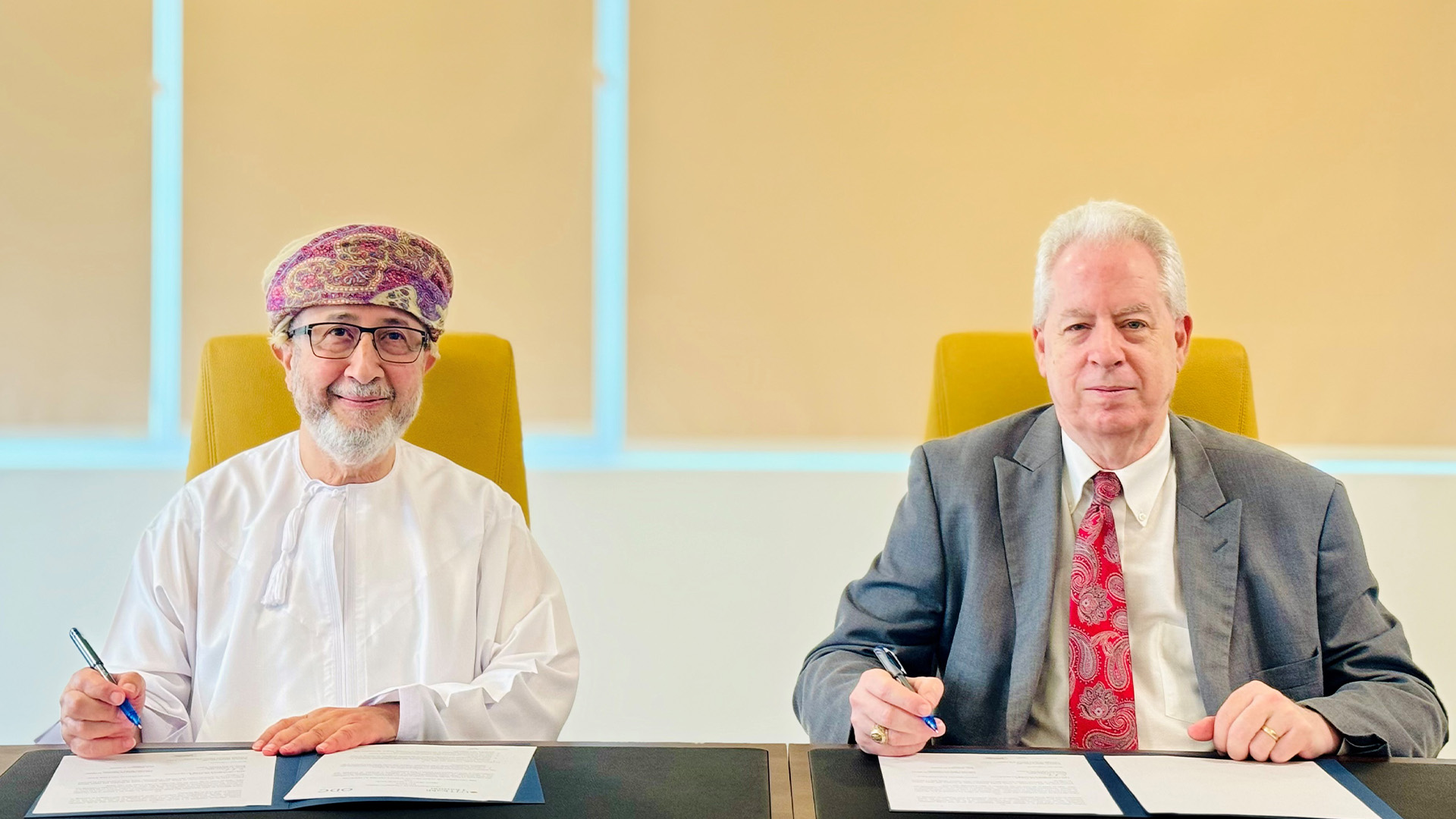 Two men seated at a table signing documents in a formal setting with beige blinds in the background.