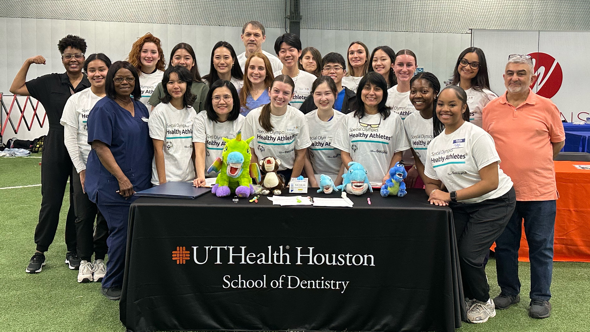 Volunteers, most wearing white “Special Olympics Healthy Athletes” shirts, pose behind a UTHealth Houston School of Dentistry table filled with dental?themed items.