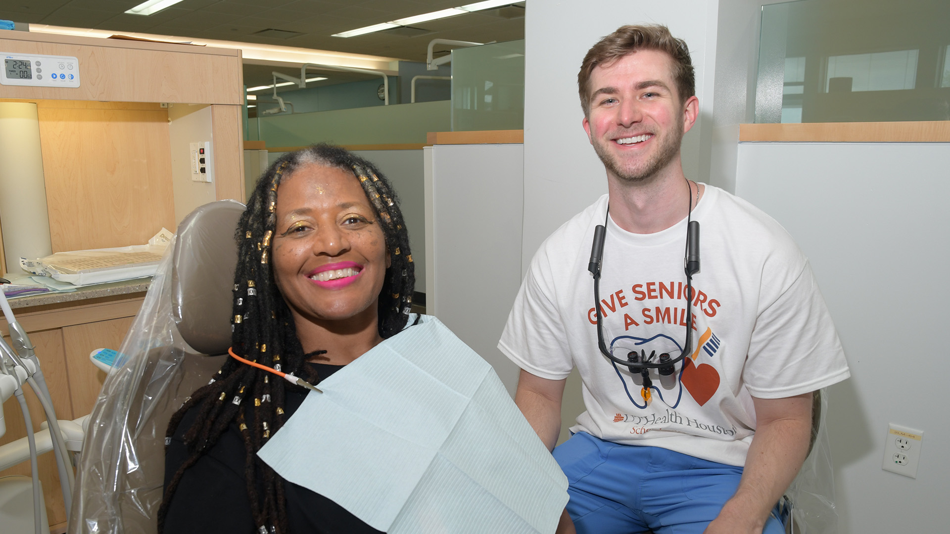 A dental student sits beside a patient in a dental chair inside a modern clinic operatory.