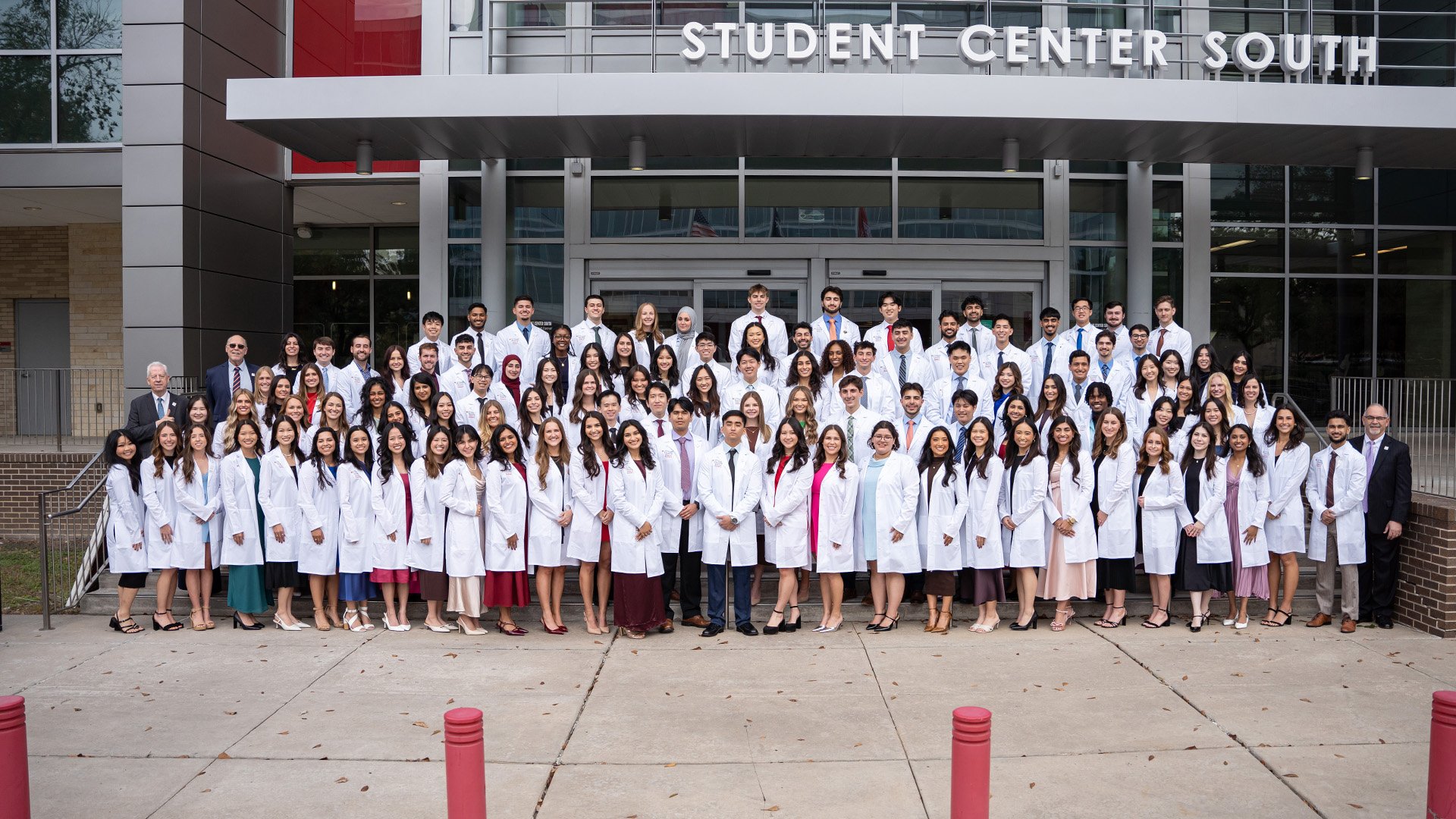 A large group of dental students stand formally wearing their white coats stand on the front steps a building for a group photo.