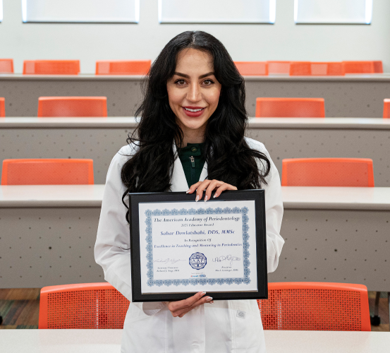Female doctor in a white laboratory coat holds an award plaque in a classroom setting.