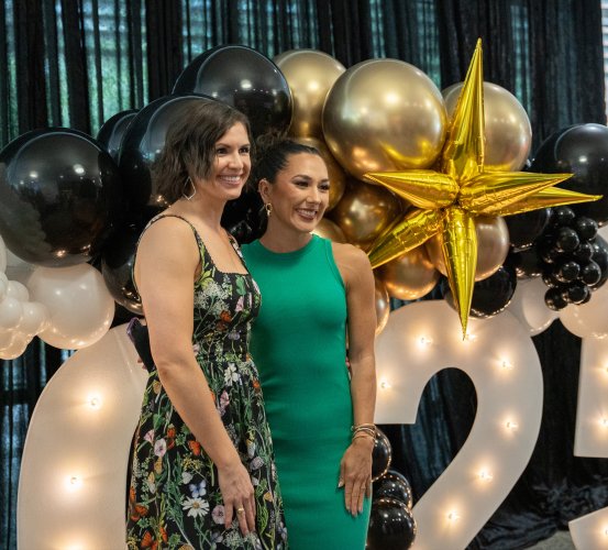 Two people standing in front of a balloon arch with white, black, and gold balloons, including a large gold star balloon.