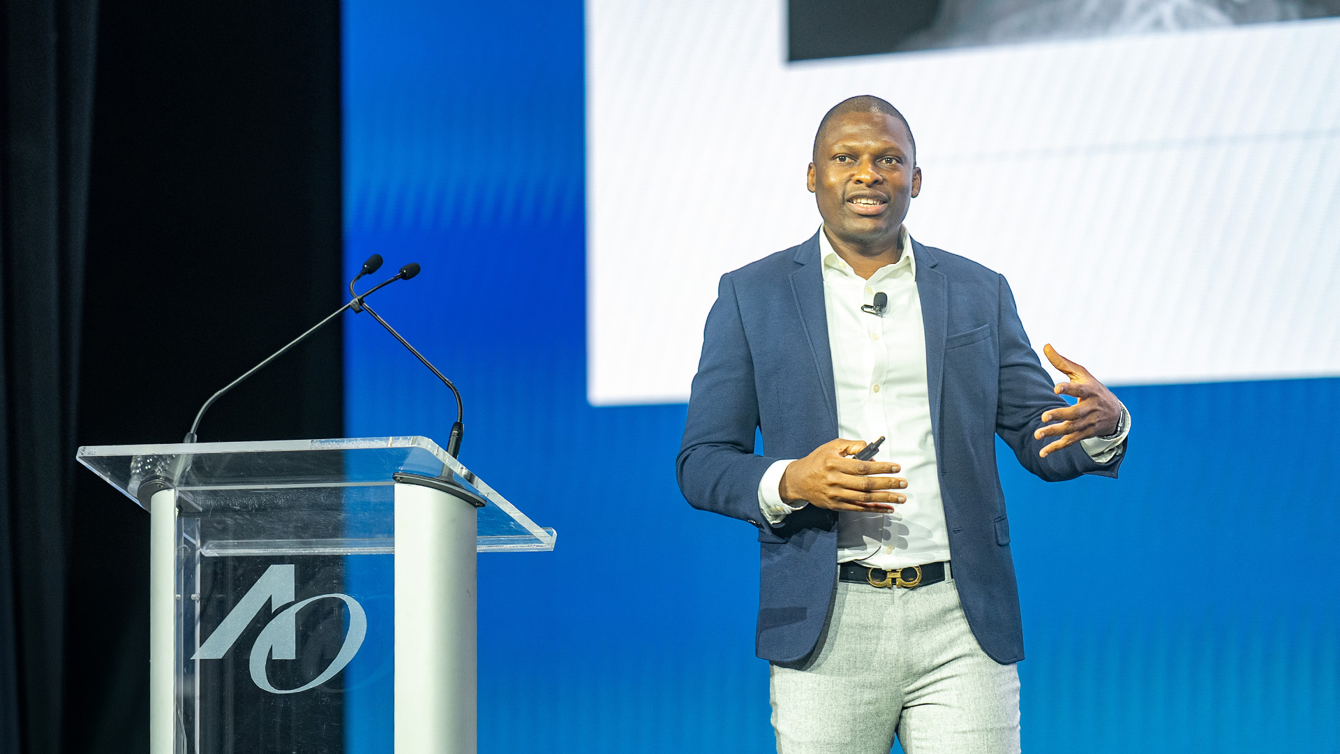 A man in business attire stands on a stage giving a presentation next to a clear podium with a stylized “AO” logo sits beside him, and a large screen behind him displaying a blue?and?white slide background.