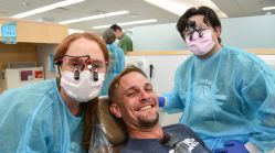 Two dental students wearing masks and protective gowns pose with a smiling patient in a dental chair during the Give Vets a Smile event at UTHealth Houston School of Dentistry.