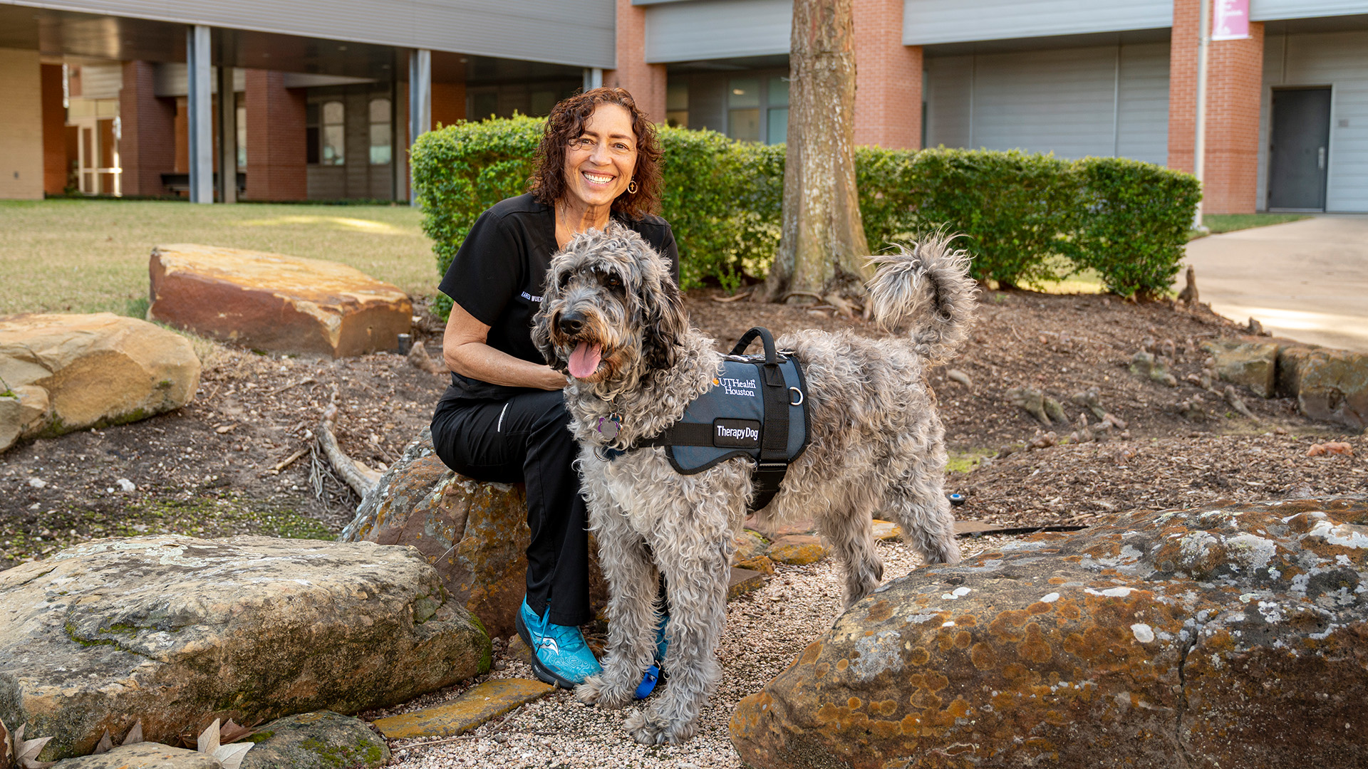 A woman sits on rocks outdoors with a gray labradoodle wearing a vest.