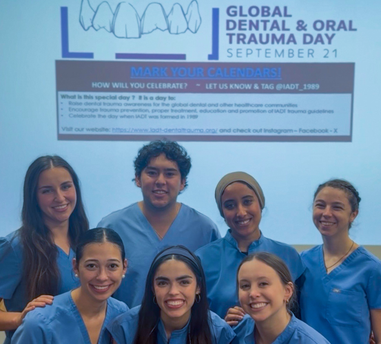 Students in light blue scrubs stand in front of a projector screen displaying information on Global Dental and Oral Trauma Day, which is observed on Sept. 21.