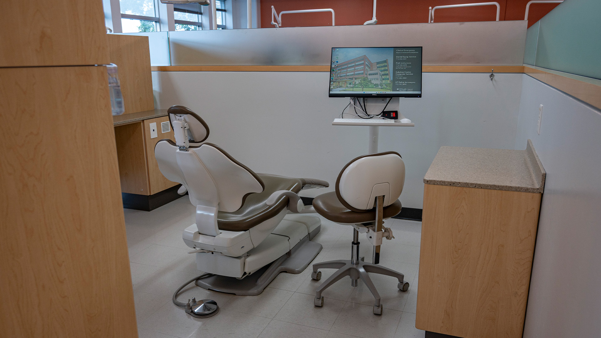 A modern dental operatory features a reclining dental chair, attached equipment, a dentist’s stool, a countertop with cabinetry, and a computer monitor, all within a bright space accented by wood paneling and frosted glass partitions.
