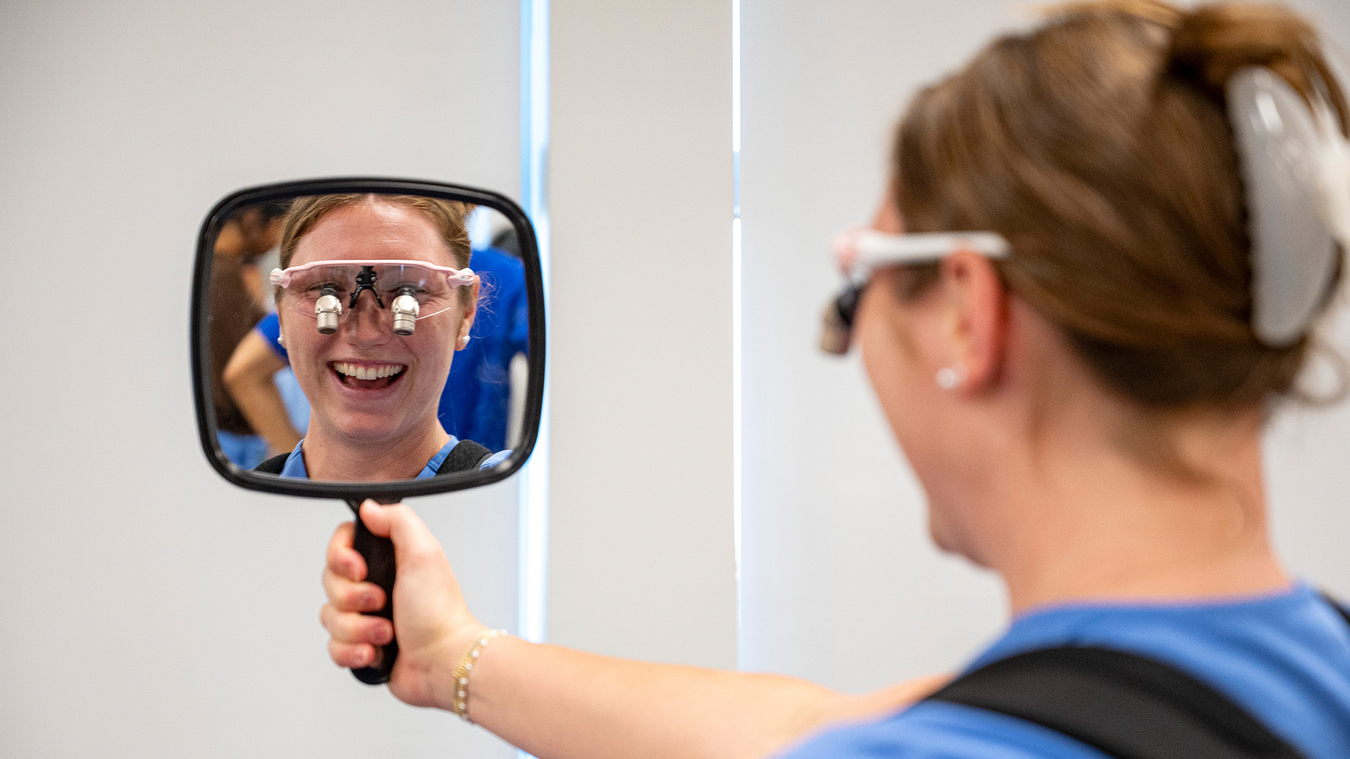 Student wearing dental loupes holds a mirror to view the reflected image of the magnification eyewear during a training session.