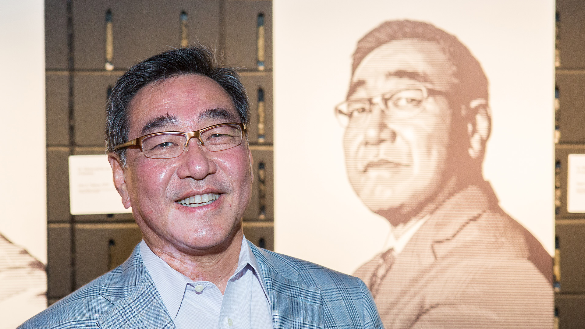 A person in a light gray plaid suit jacket stands in front of a large stylized portrait depicting the same individual, set against a wall with dark panels and small labels.