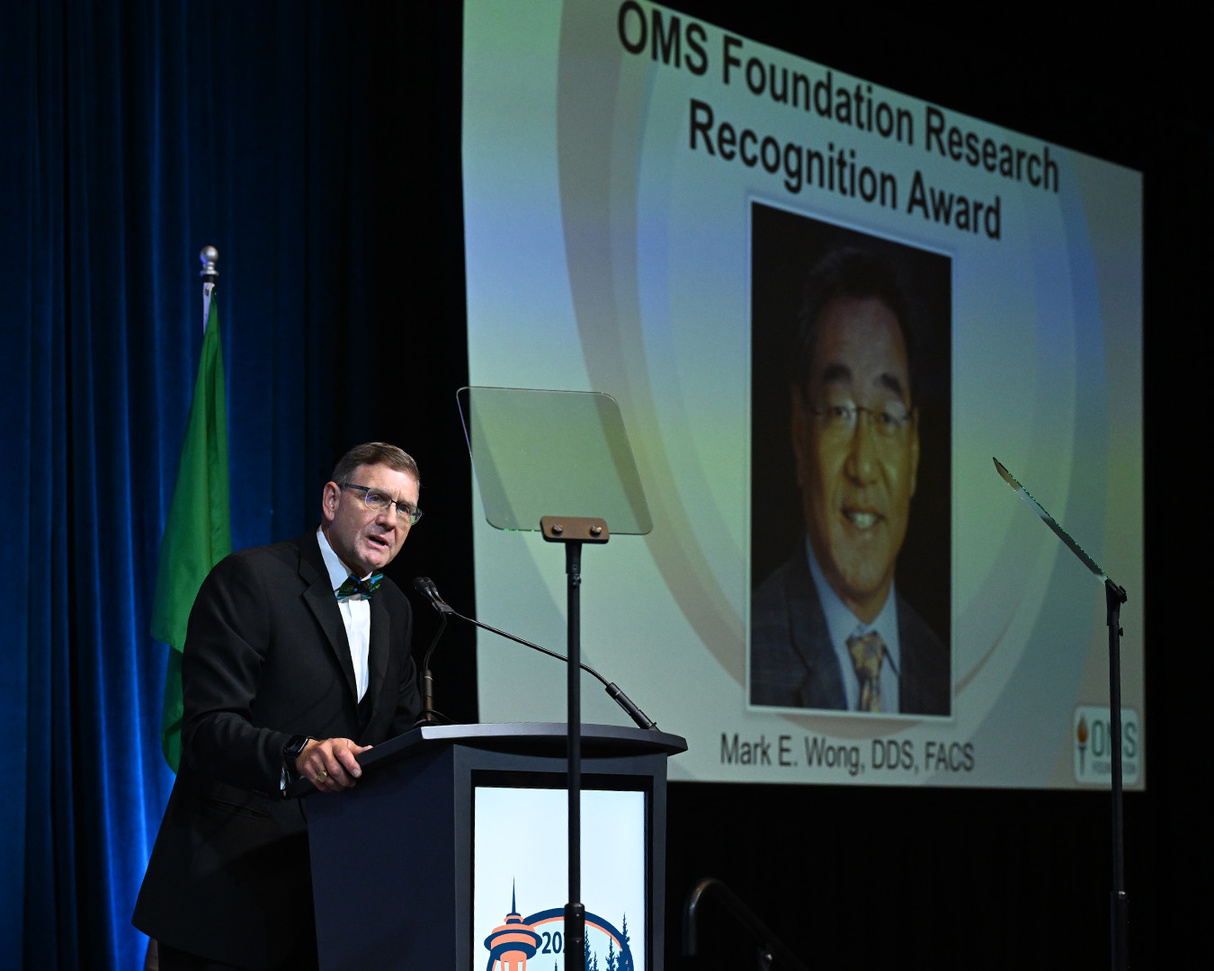 A speaker stands at a podium during an awards program while a large screen behind displays an OMS Foundation Research Recognition Award presentation slide featuring a recipient's photo and award title.