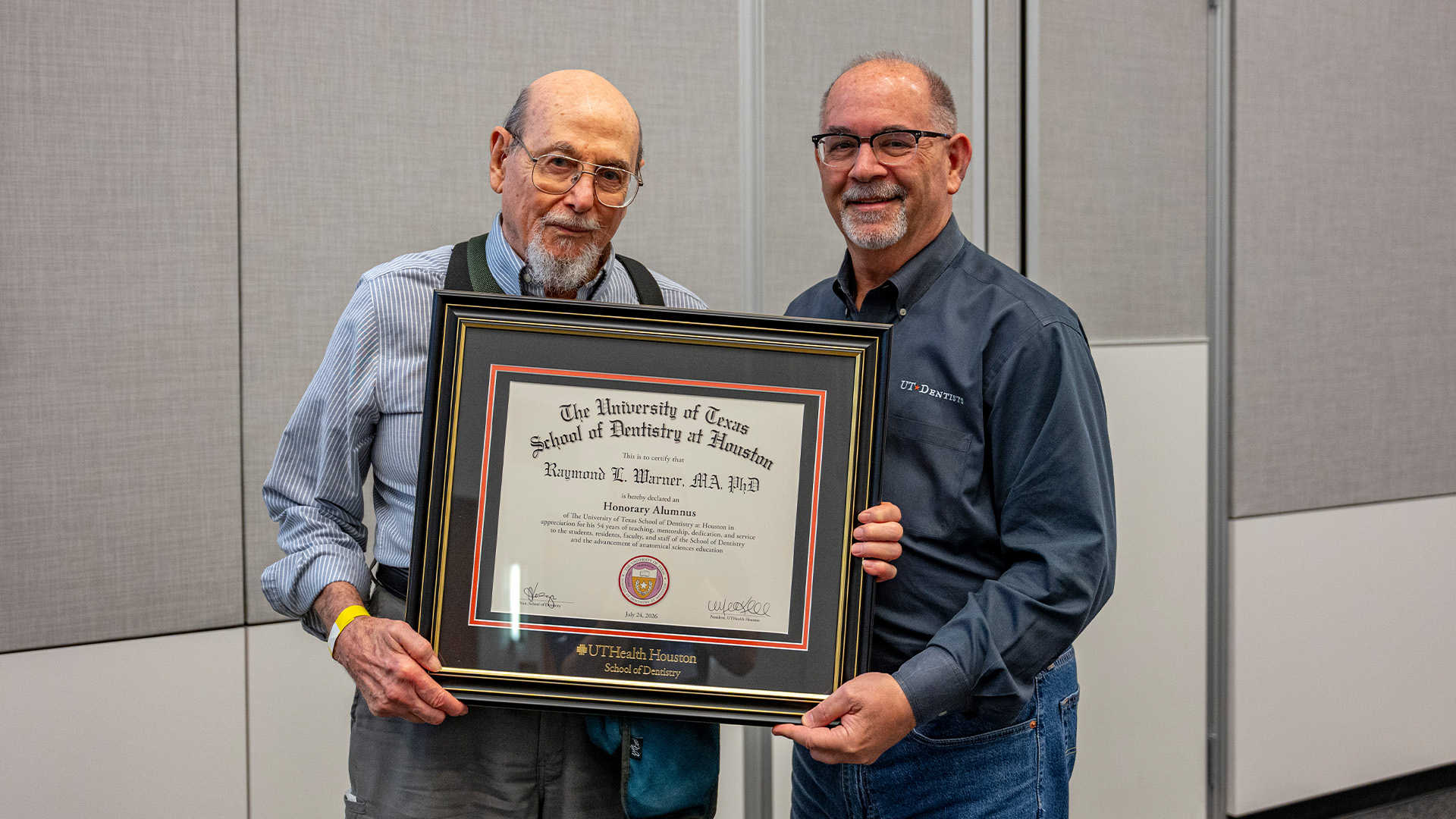 Two individuals stand indoors holiding a framed UTHealth Houston School of Dentistry certificate recognizing Raymond L. Warner as an Honorary Alumnus