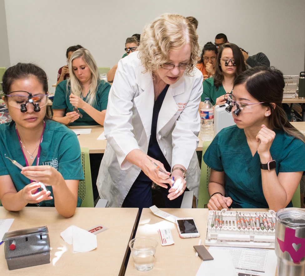 At a table in a classroom, Clinical Assistant Professor Ruth Conn, RDH, MSDH, shows a dental hygiene student how to sharpen and instrument.