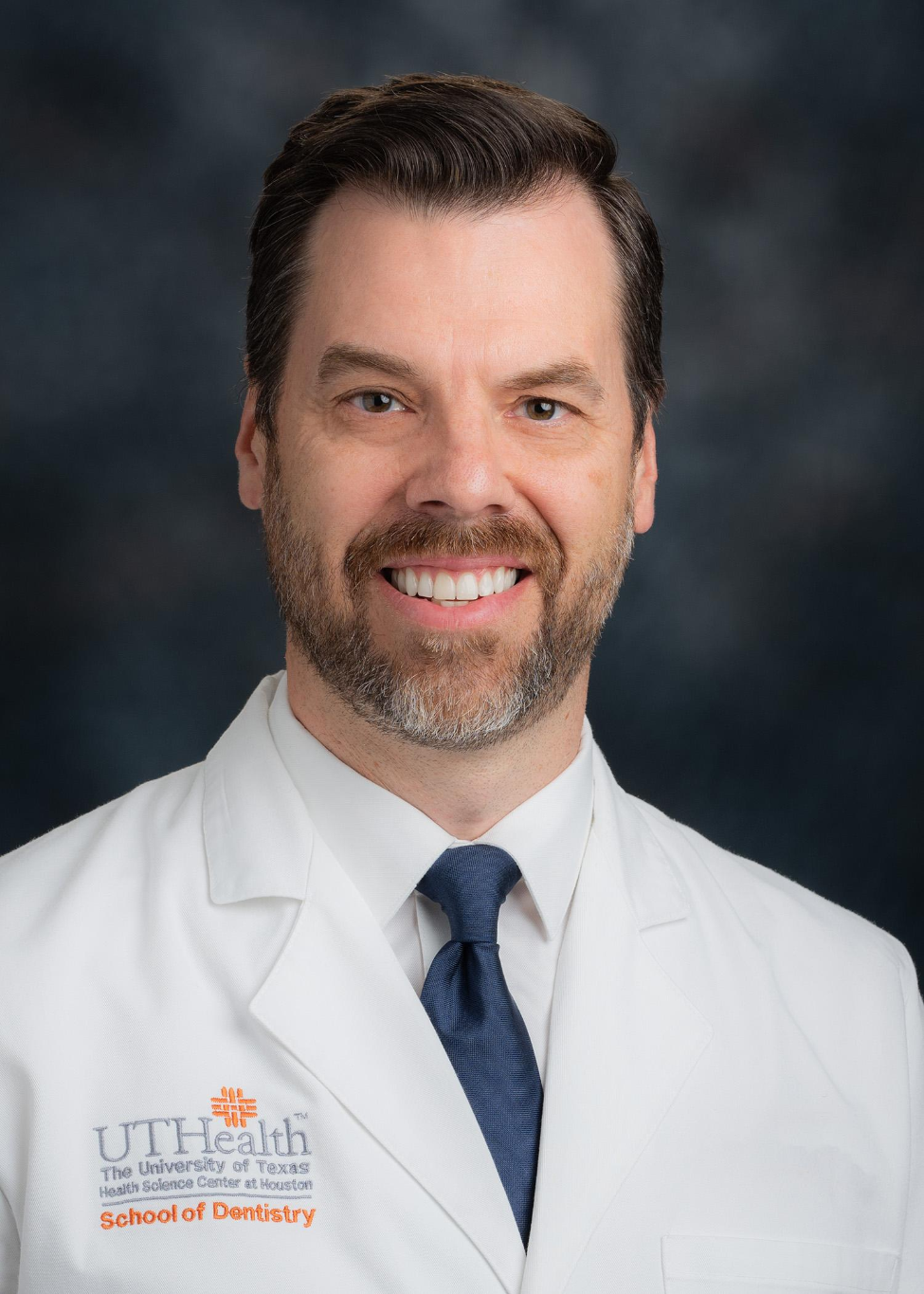 Dr. Greg Olson wearing a white coat and tie. The coat has the UTHealth School of Dentistry logo.