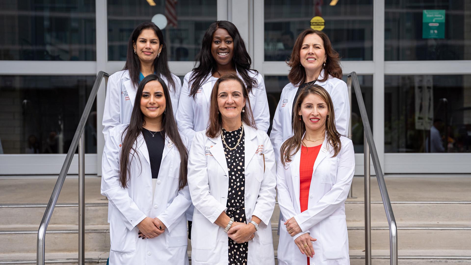 Six women stand on the steps outside a building entrance, all wearing white coats. They are arranged in two rows of three and are posed formally.