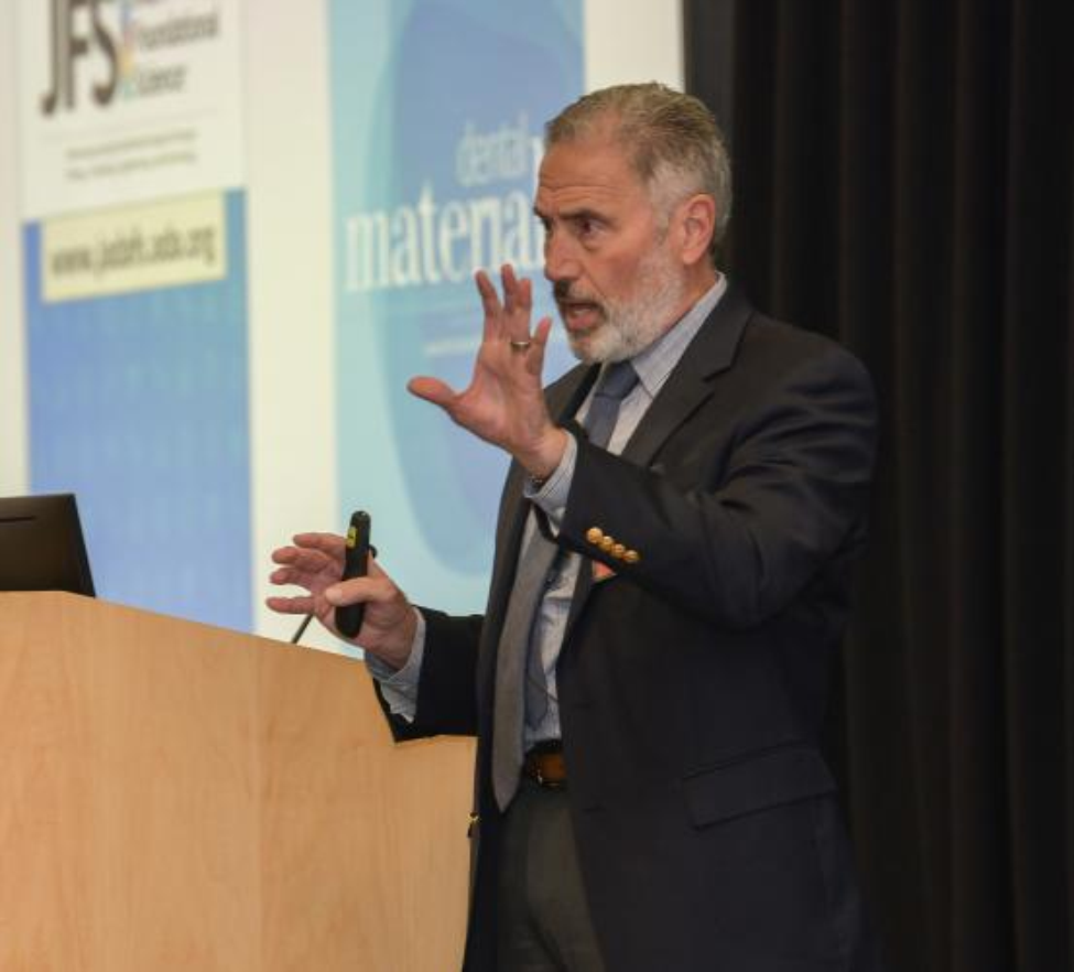 Man in a suit stands next to a podium presenting.