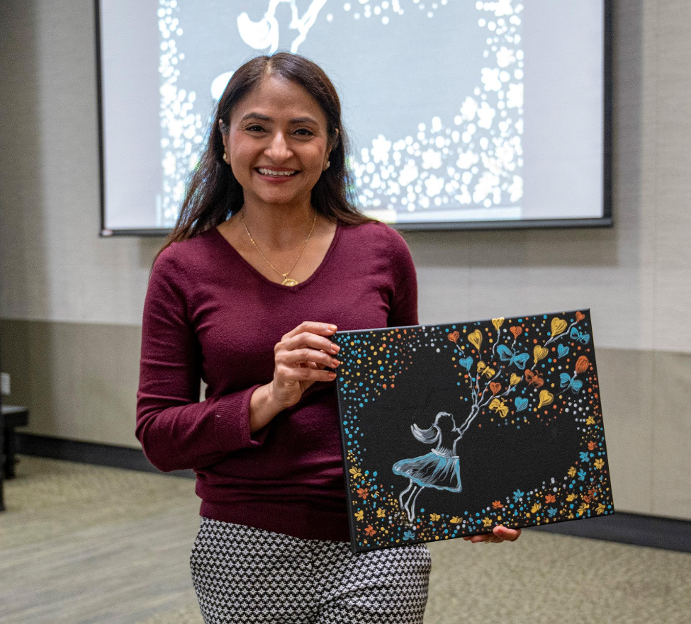 A woman stands indoors, smiling and holding a piece of artwork. The artwork depicts a stylized girl in a dress being lifted by colorful balloons against a black background with scattered dots resembling stars.