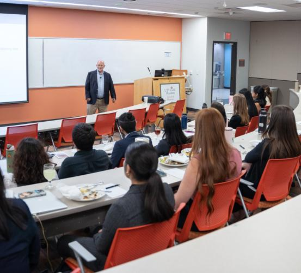 A man in a navy blazer speaks to students seated in a classroom during a luncheon lecture.