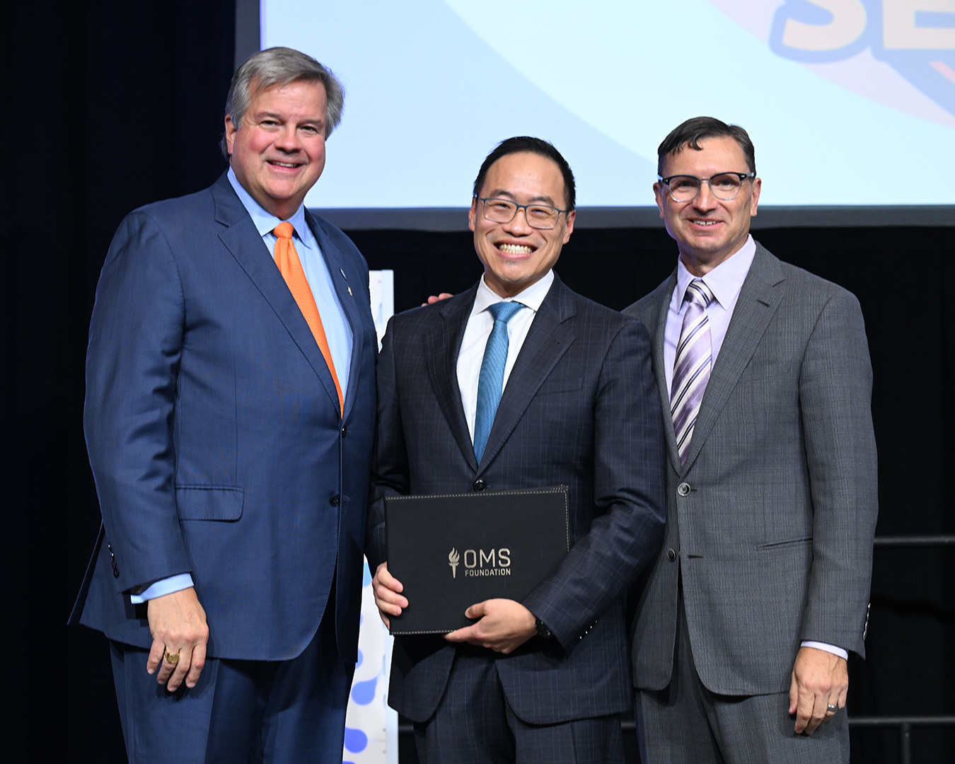 Three individuals stand onstage at an awards event. The person in the center holds an OMS Foundation certificate folder while posing with two others in business attire in front of a presentation screen.