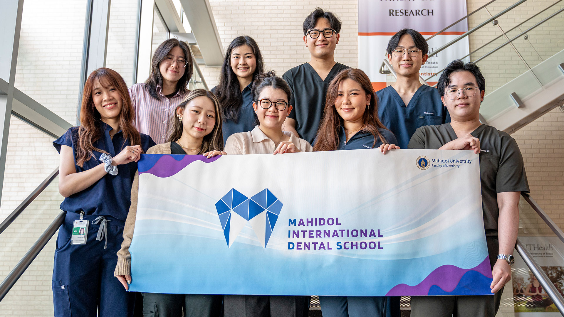 Group of students and trainees on a staircase holding a Mahidol International Dental School banner inside a university building.