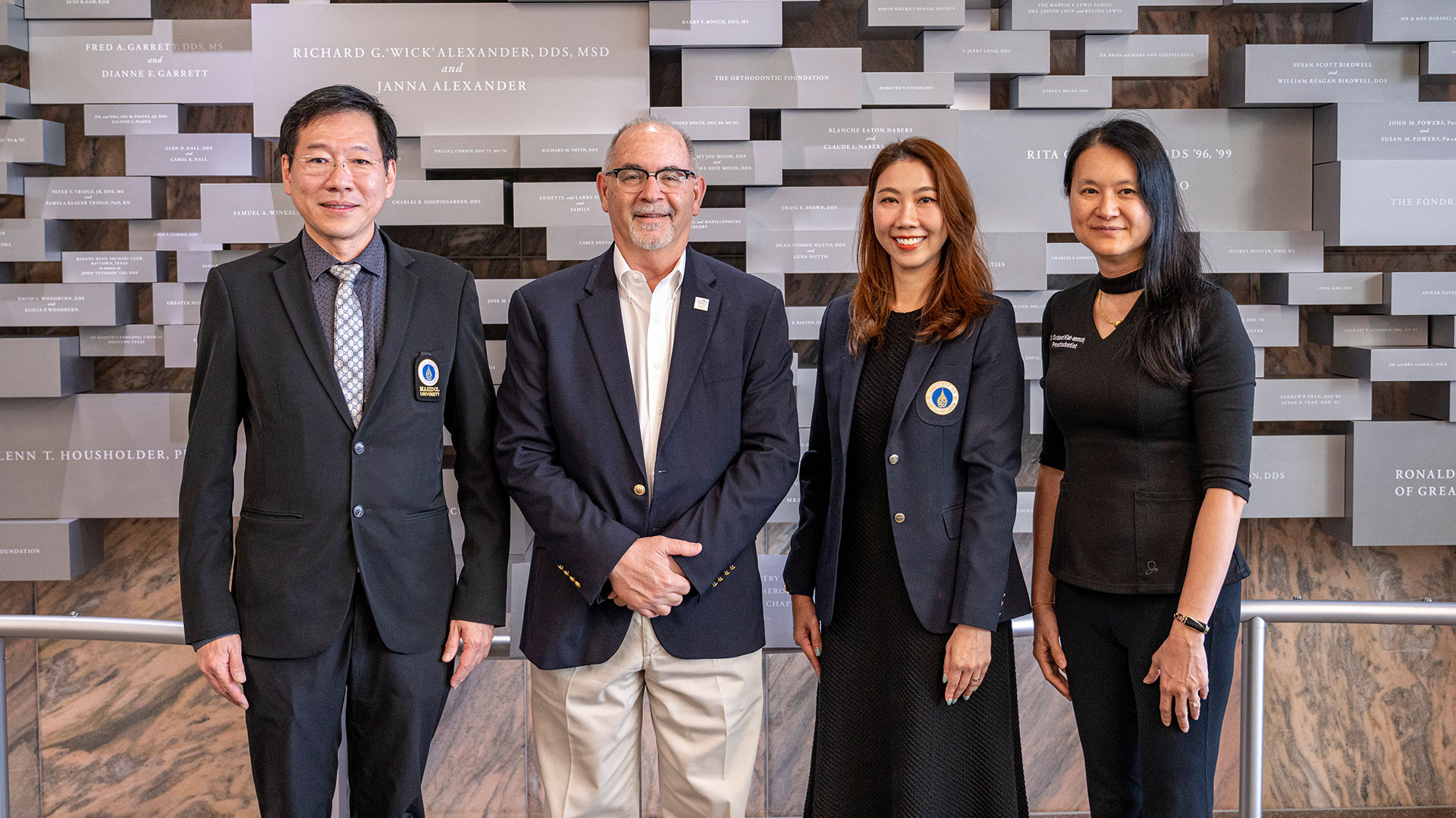 Four visitors pose in front of a donor recognition wall inside a dental school building.