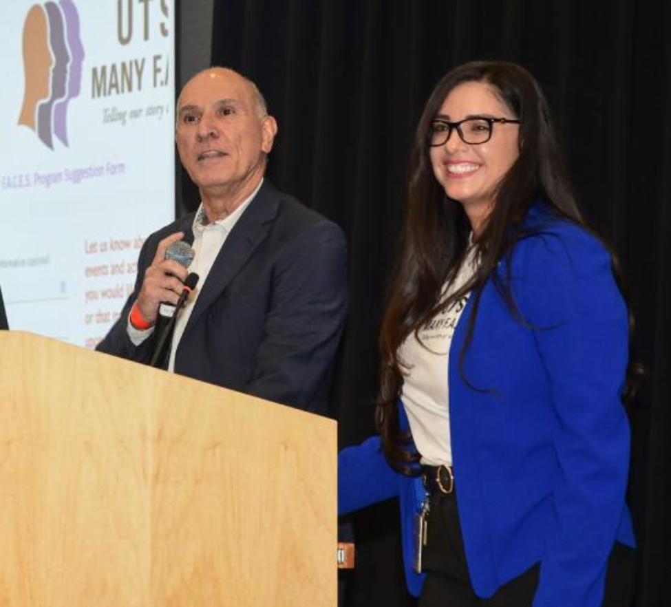 Two people in professional dress stand presenting at a podium with a projector screen in the background.