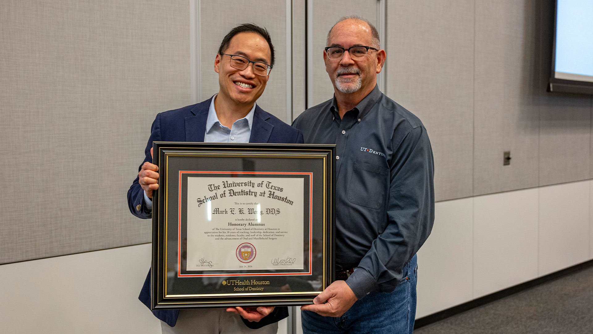 Two individuals stand indoors holiding a framed UTHealth Houston School of Dentistry certificate recognizing Mark E. K. Wong, as an Honorary Alumnus