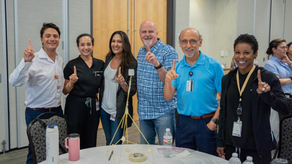 Six smiling faculty members celebrate winning the Marshmallow Challenge, posing with their spaghetti and marshmallow tower. The team holds up one finger and proudly gathers around the table during the team-building activity.