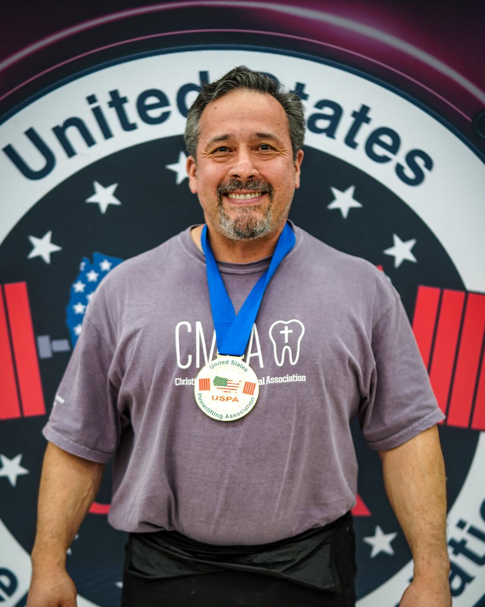 Man poses for picture at powerlifting competition while wearing first-place medal.