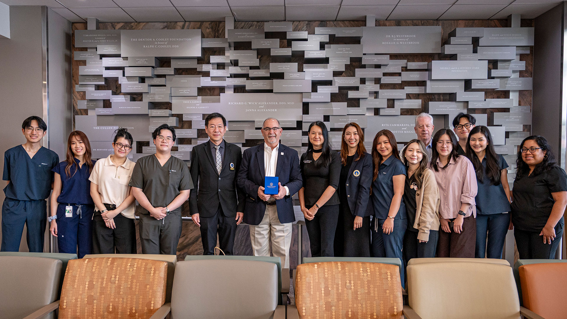 Group photo of students, faculty, and visitors standing in front of a donor recognition wall inside a dental school building. One person at the center holds a blue commemorative plaque.