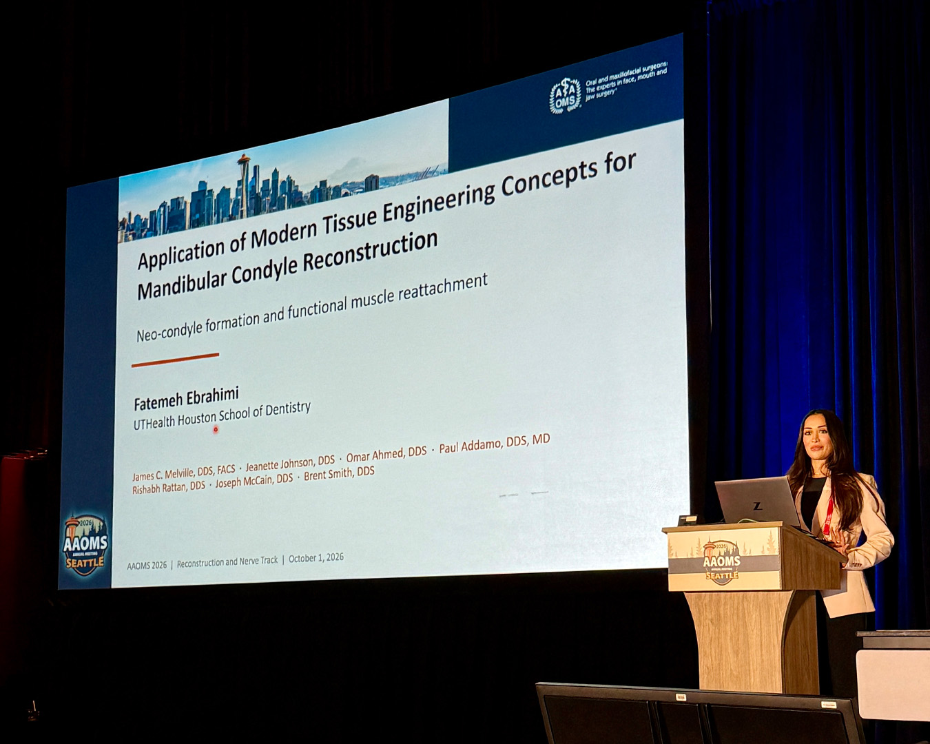 A presenter stands at a podium beside a large projection screen displaying a research presentation at a professional conference. The slide covers tissue engineering concepts for mandibular condyle reconstruction.