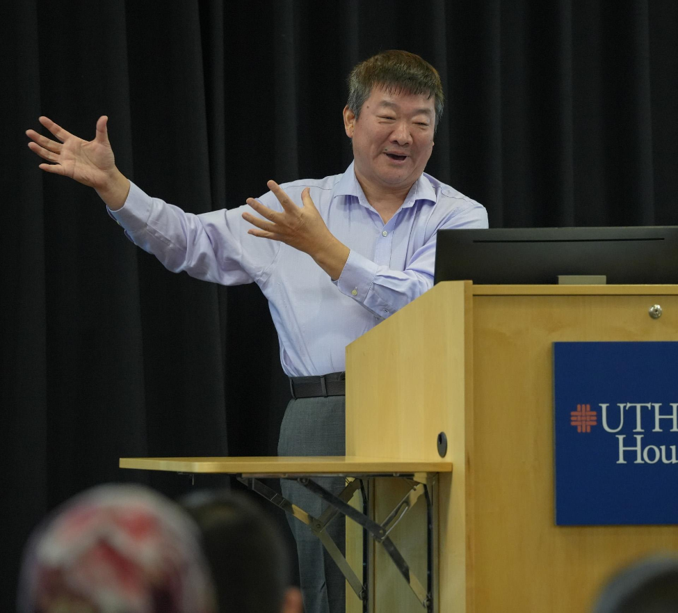 Person giving a presentation at a podium with a UTHealth Houston sign, gesturing with both hands.