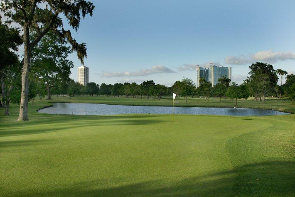 View of one of the greens at Hermann Park Golf Course with a water hazard and oak trees in the background.