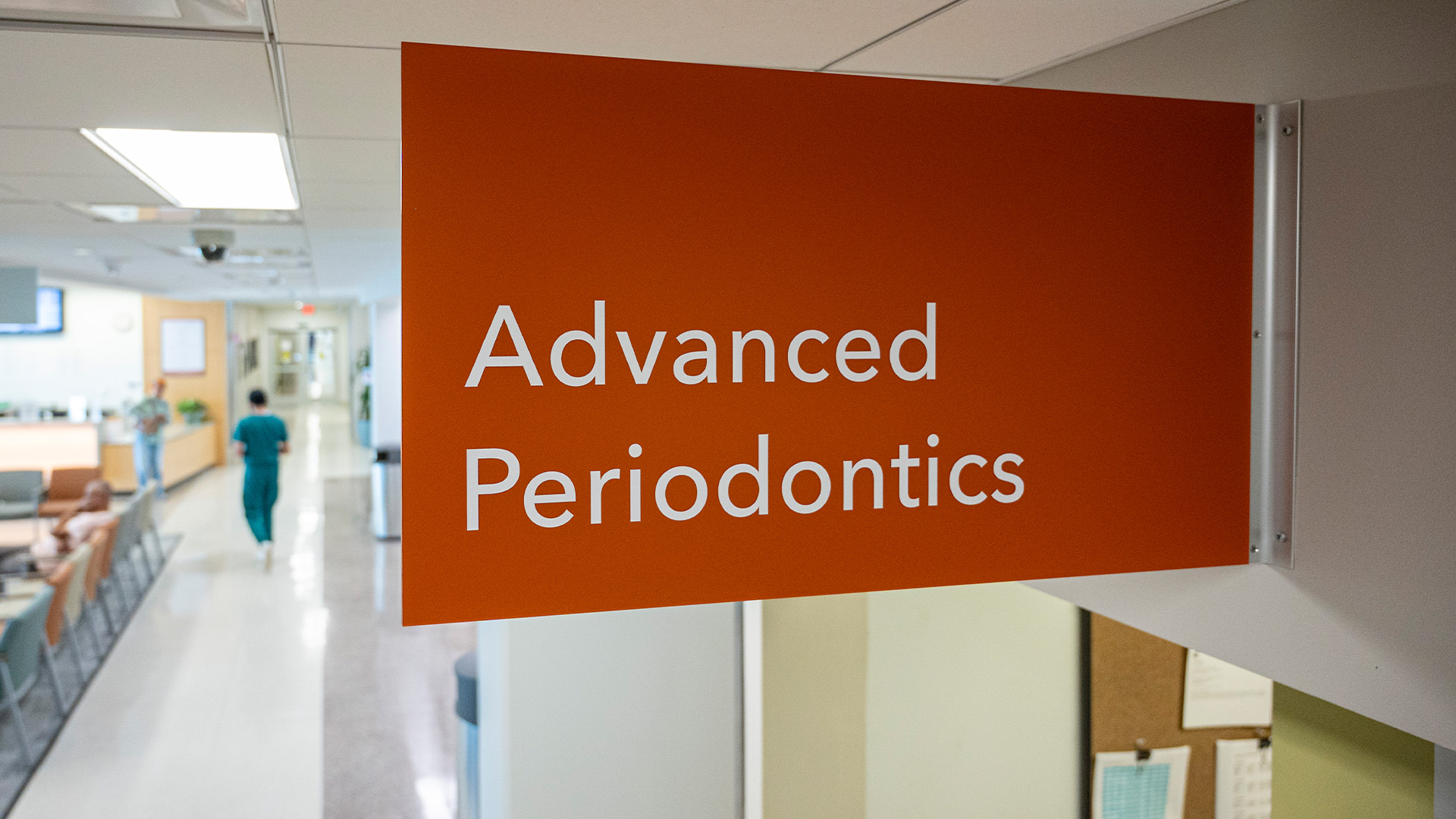 A hallway inside a clinical facility features a large orange wall sign that reads “Advanced Periodontics” in white letters. In the background, people in scrubs and casual clothing are visible near a waiting area with chairs and a reception desk.