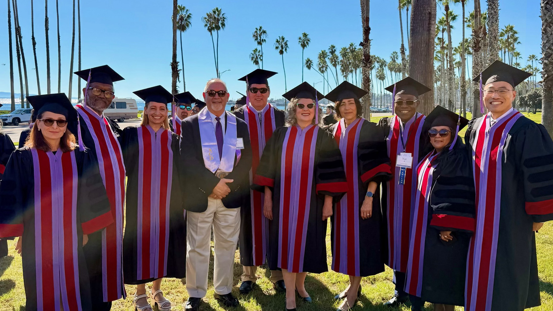 A group in graduation regalia gathers for a photo outdoors beneath a clear blue sky. Rows of palm trees and a waterfront setting form the backdrop.