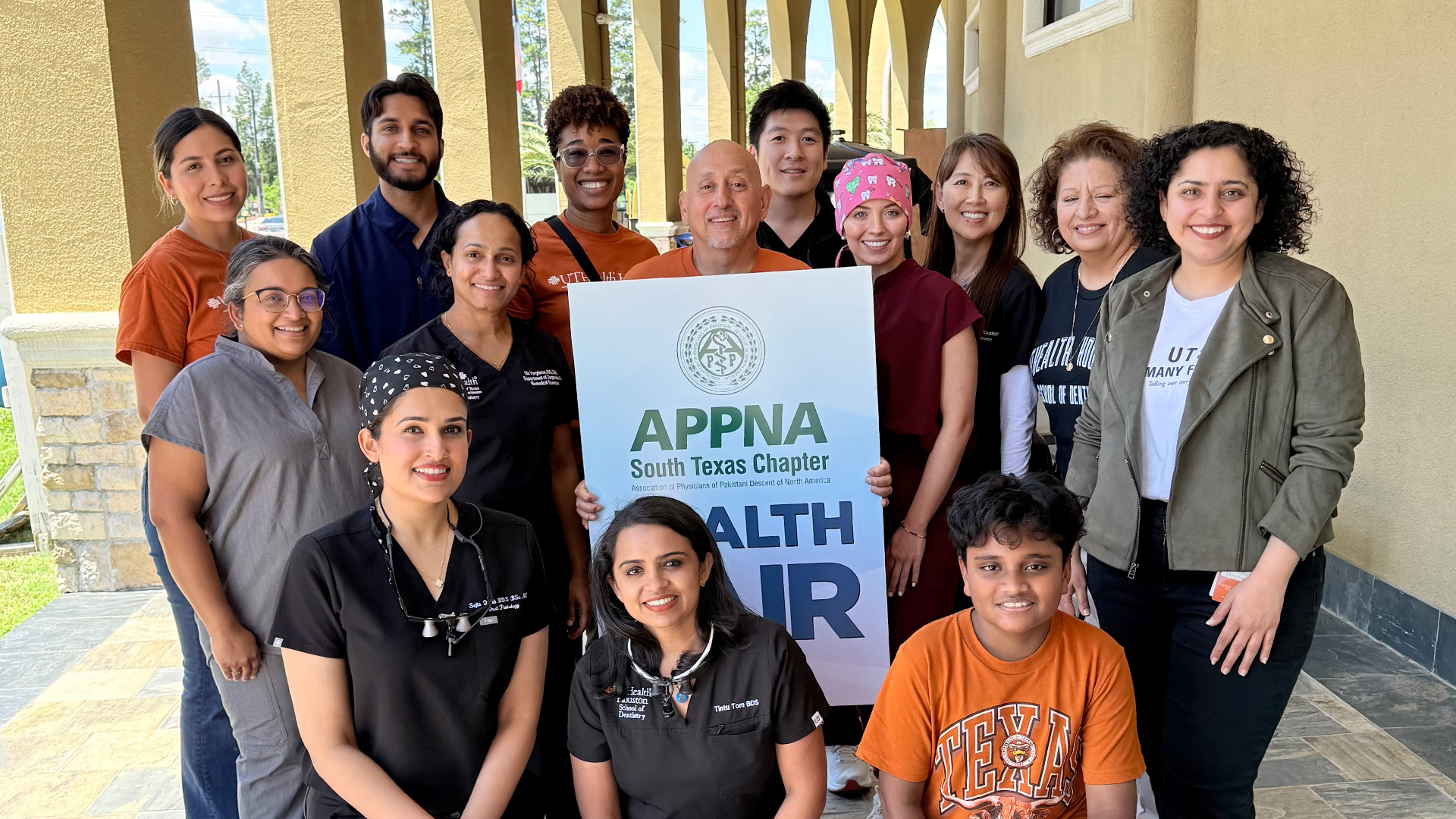 Fourteen people pose together outdoors under a covered walkway with tall arches and a tiled floor. They stand and kneel around a sign that reads “APPNA South Texas Chapter Health Fair.”