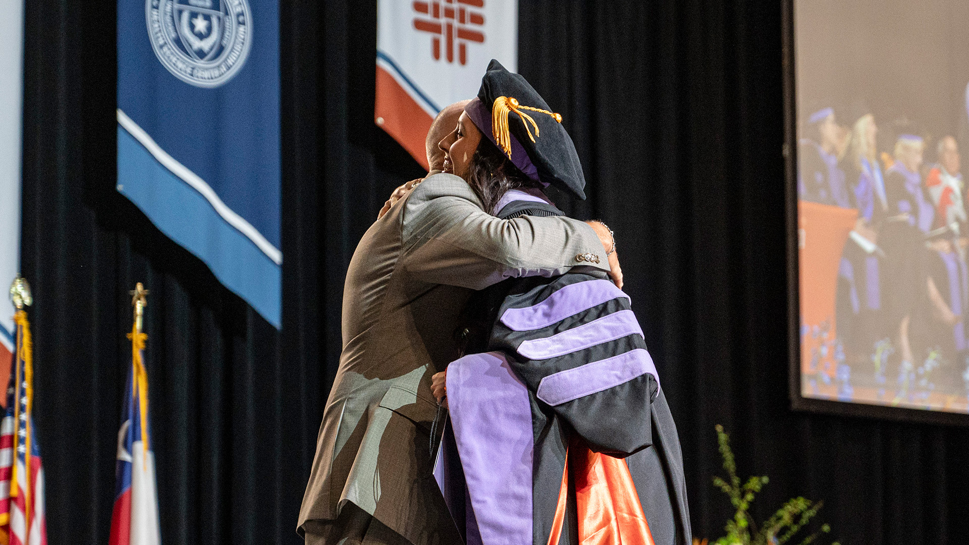A female graduate in black doctoral regalia with purple and orange accents hugs a male family member on stage during a commencement ceremony. Behind them are university banners, the Texas and U.S. flags, and a large screen showing a live feed of the event.