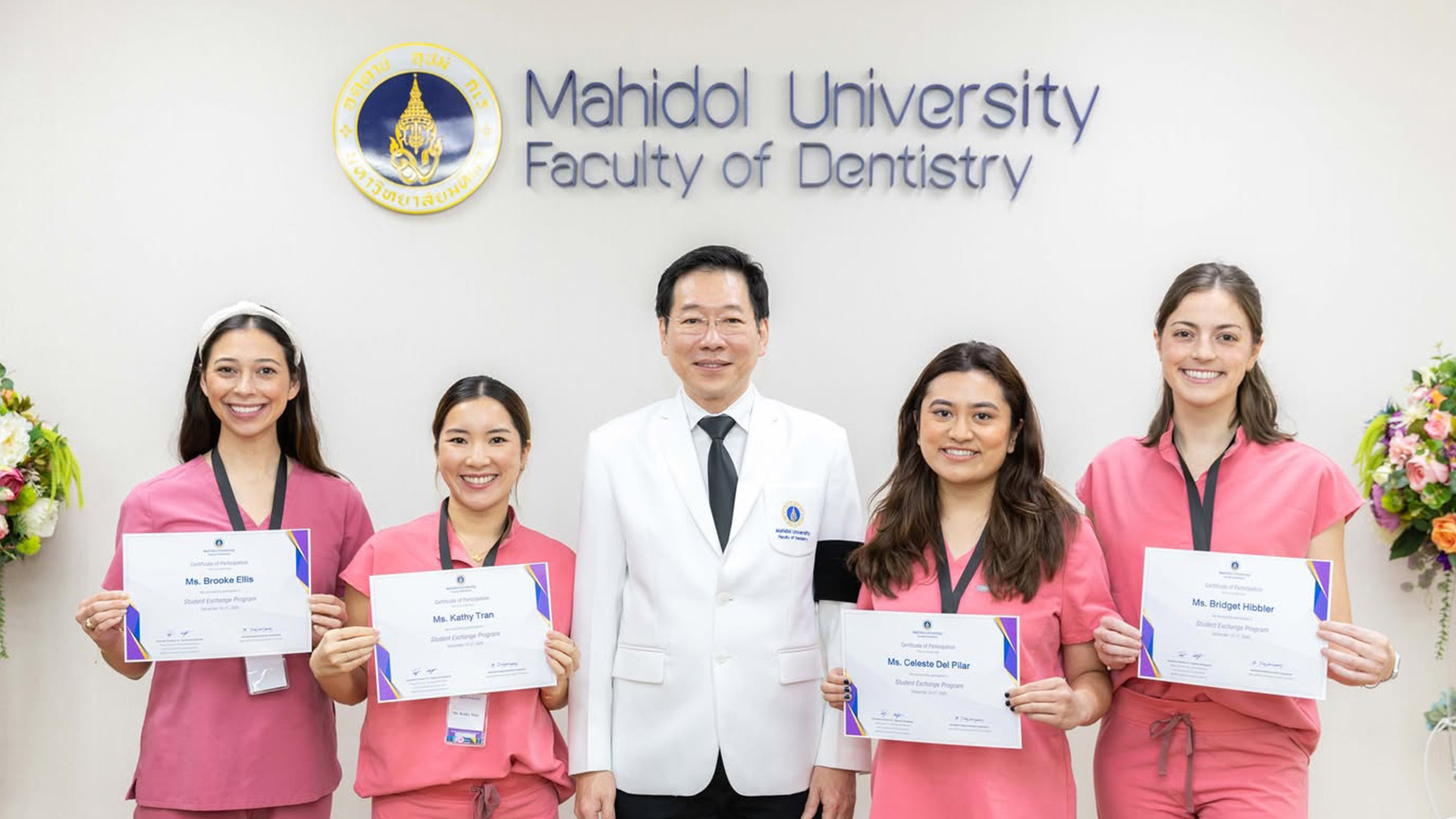 Five individuals stand in front of a wall labeled “Mahidol University Faculty of Dentistry.” Four participants in pink scrubs hold certificates from a student exchange program, while a person in a white coat stands centered with them.