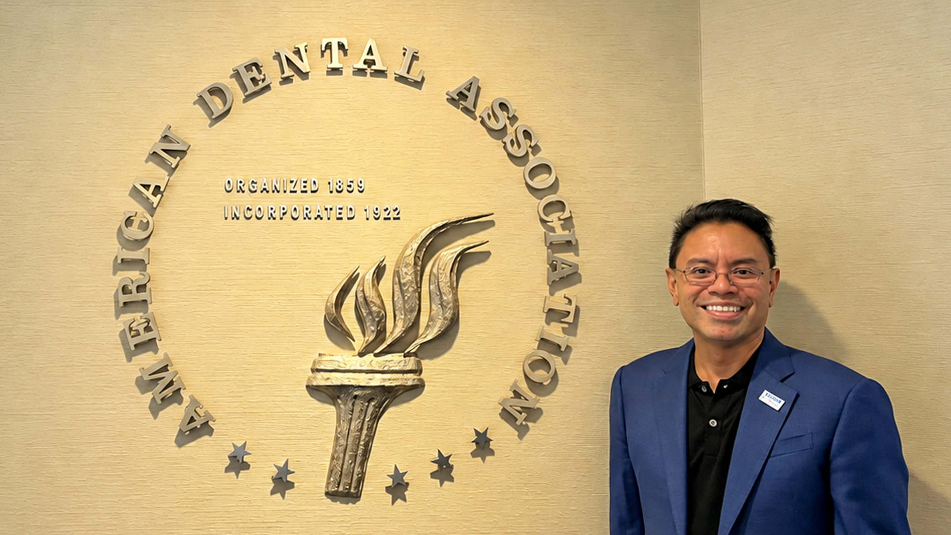 A person in a blue blazer poses next to an American Dental Association wall display featuring the association’s torch emblem and founding dates.