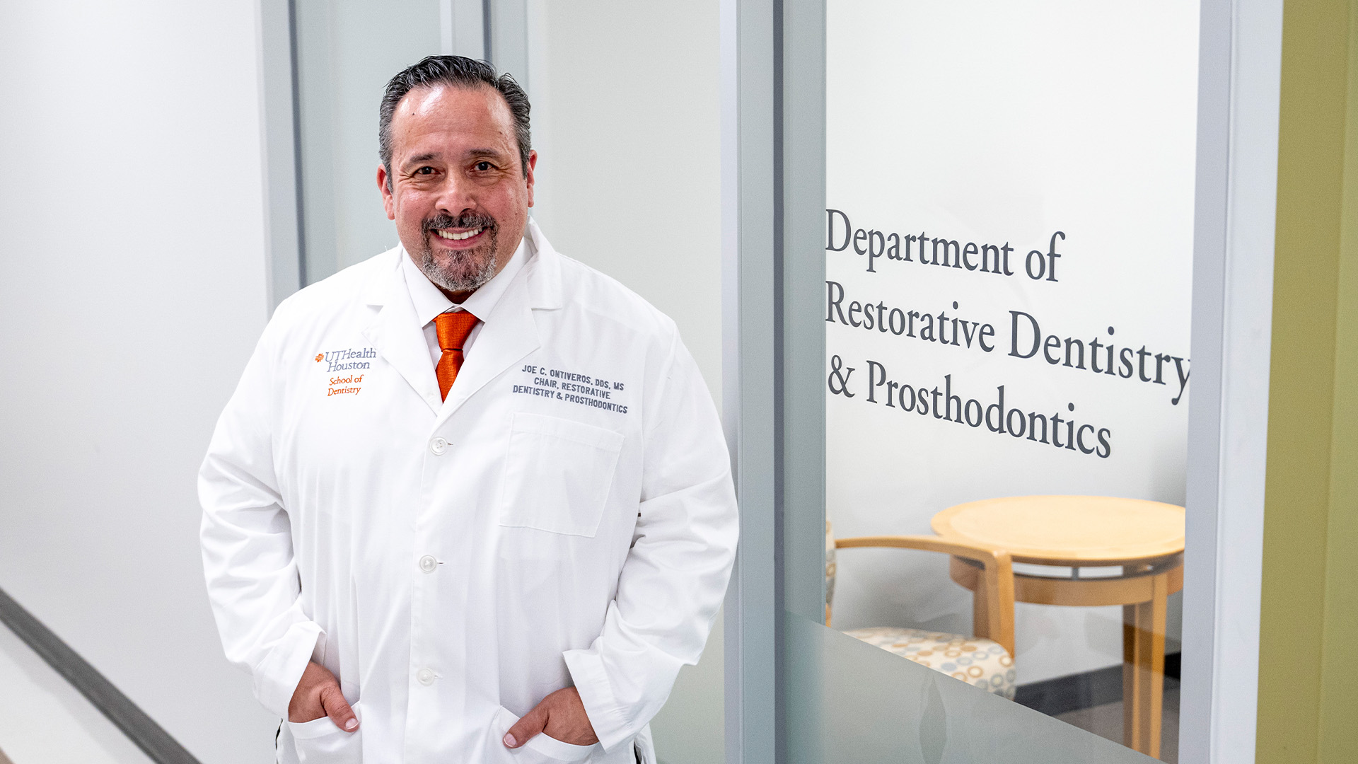 A person in a lab coat stands in a hallway outside the Department of Restorative Dentistry & Prosthodontics. A UTHealth Houston School of Dentistry logo is visible on the coat.