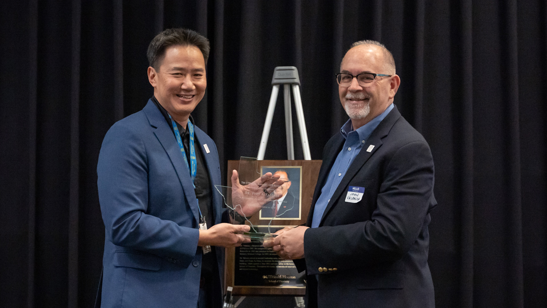 Two people in business attire stand in front of a dark curtain, smiling as they hold a transparent glass award shaped like the state of Texas. An easel beside them displays a framed plaque with a photo and text.