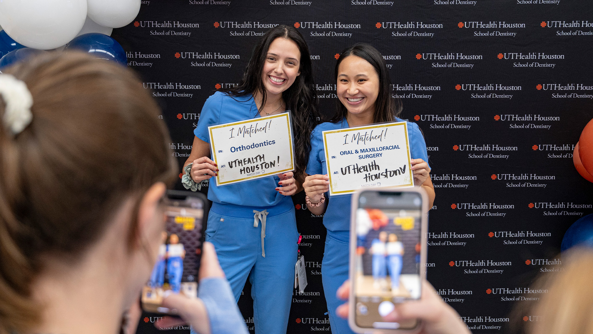 Two people in blue scrubs hold residency match signs as others photograph them.